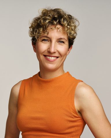 Mujer sonriente con cabello rizado, vistiendo una blusa naranja. Fondo neutro.