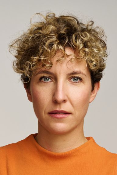 Persona con cabello rizado y corto, vistiendo una camiseta naranja, mirada seria.